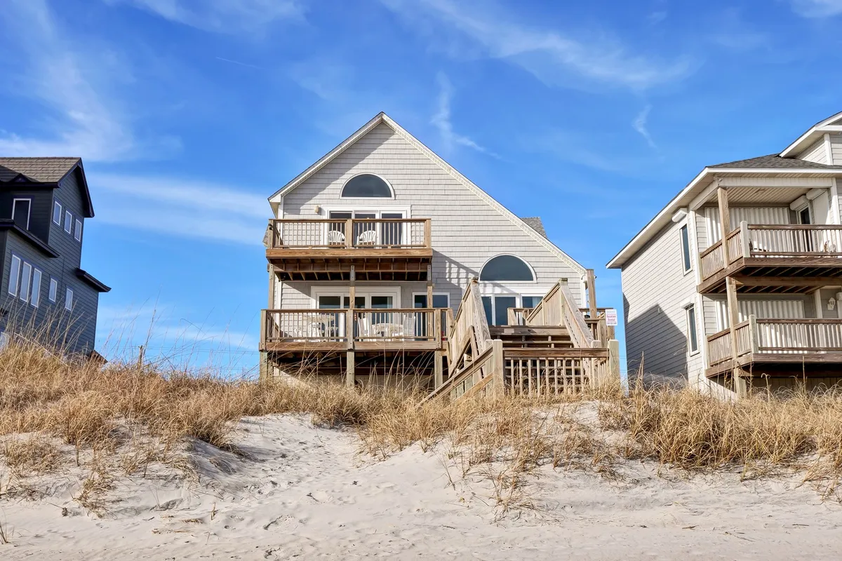 A charming beach house with spacious balconies overlooks sand dunes, set against the backdrop of the wide ocean. The open decks hint at spaces for adults to unwind and families to gather, enjoying ocean breezes and sunset views together.