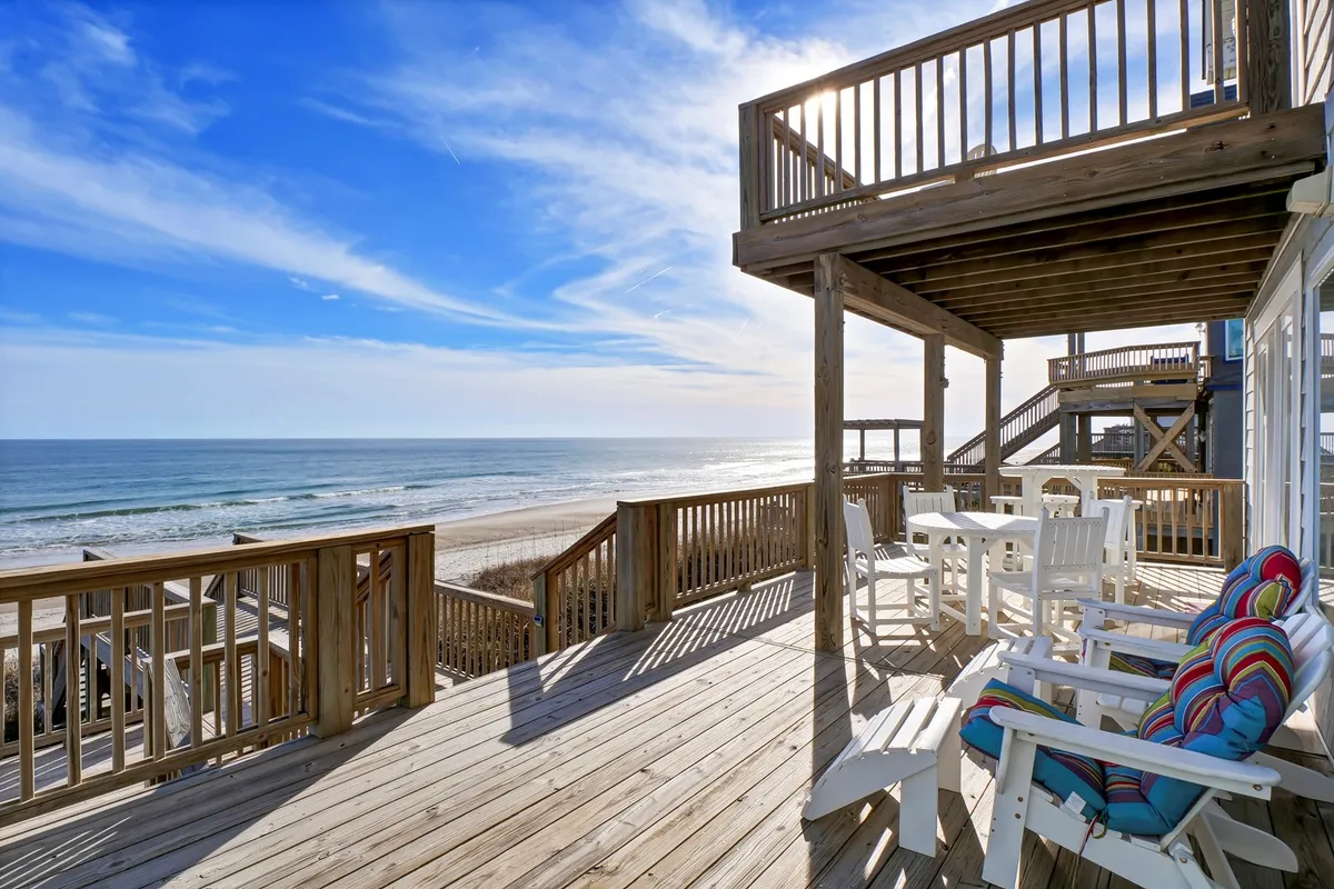 An airy wooden deck furnished with comfortable seating and a dining area, all overlooking a stunning ocean vista. Wooden railings frame the view, inviting guests to move effortlessly from indoor comfort to sun-soaked outdoor lounging.