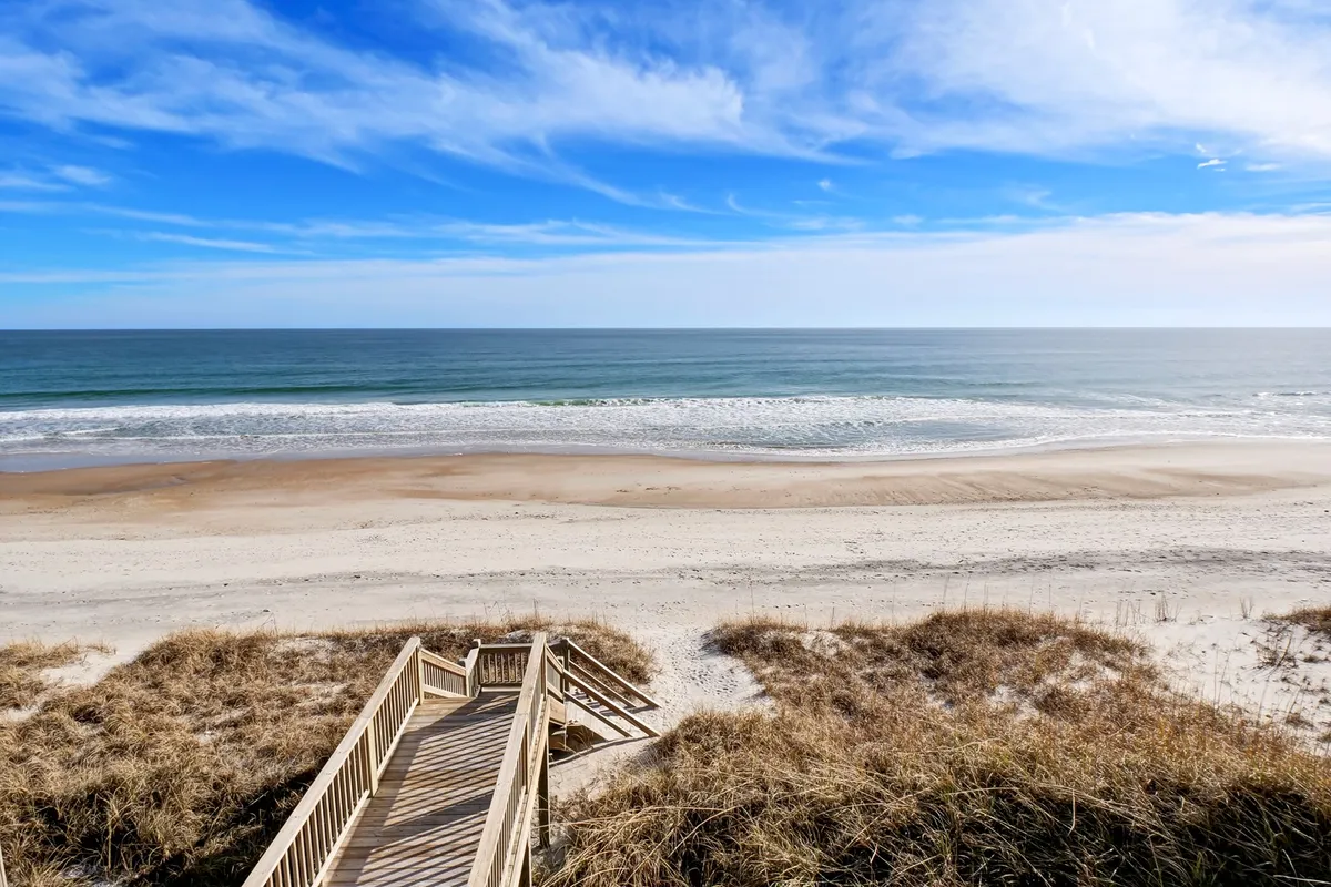 A serene wooden boardwalk stretches over dune grass, leading directly to a sunlit sandy beach where gentle waves roll in. The scene evokes the excitement of children running toward the water, ready for sandcastle-building and splashing in the surf.