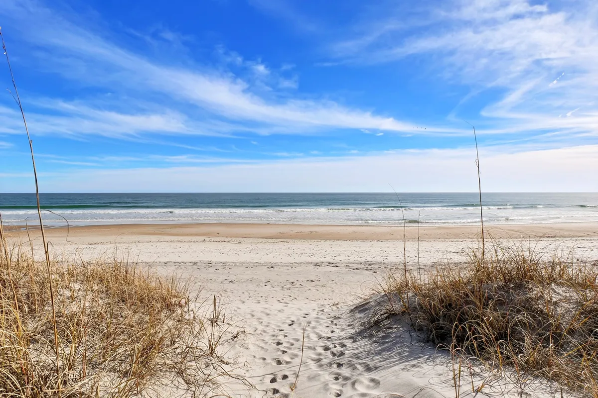 A coastal landscape featuring sand dunes, gently breaking ocean waves, and a wide blue sky. The untouched beach invites exploration, water sports, or sunset strolls, highlighting the natural beauty of the surroundings.