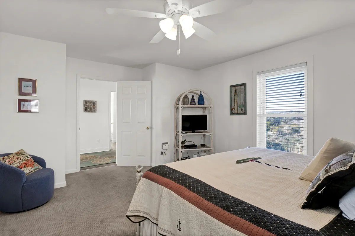 A cozy queen bedroom with a quilted bed, blue armchair, and a shelf with decorative accents. The room is filled with morning sunlight, offering a peaceful space perfect for adults or children. Inviting and serene, the design balances comfort and versatility.