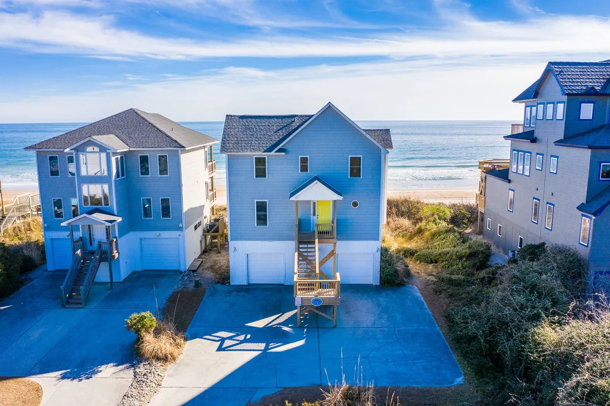 A beachfront property with a blue exterior, a spacious wooden deck overlooking the water, and surrounding vegetation. The deck offers views perfect for sunrise coffee, sunset cocktails, or peaceful relaxation by the sea.
