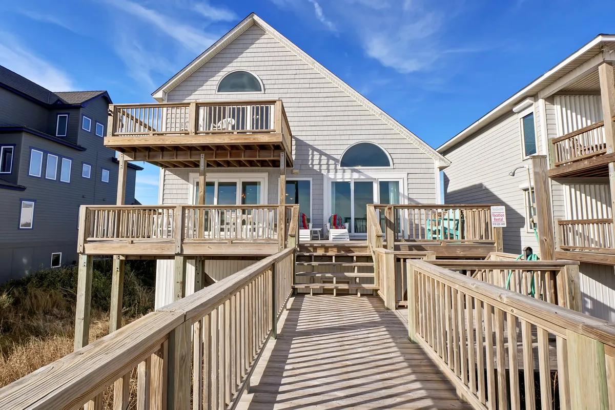 A relaxed exterior scene featuring a wooden deck and balcony with ample seating, large windows, and views of surrounding buildings. The deck is ideal for outdoor dining, sunbathing, or simply enjoying the sea air with friends and family, perfectly suited for sunset dinners or morning coffee together.