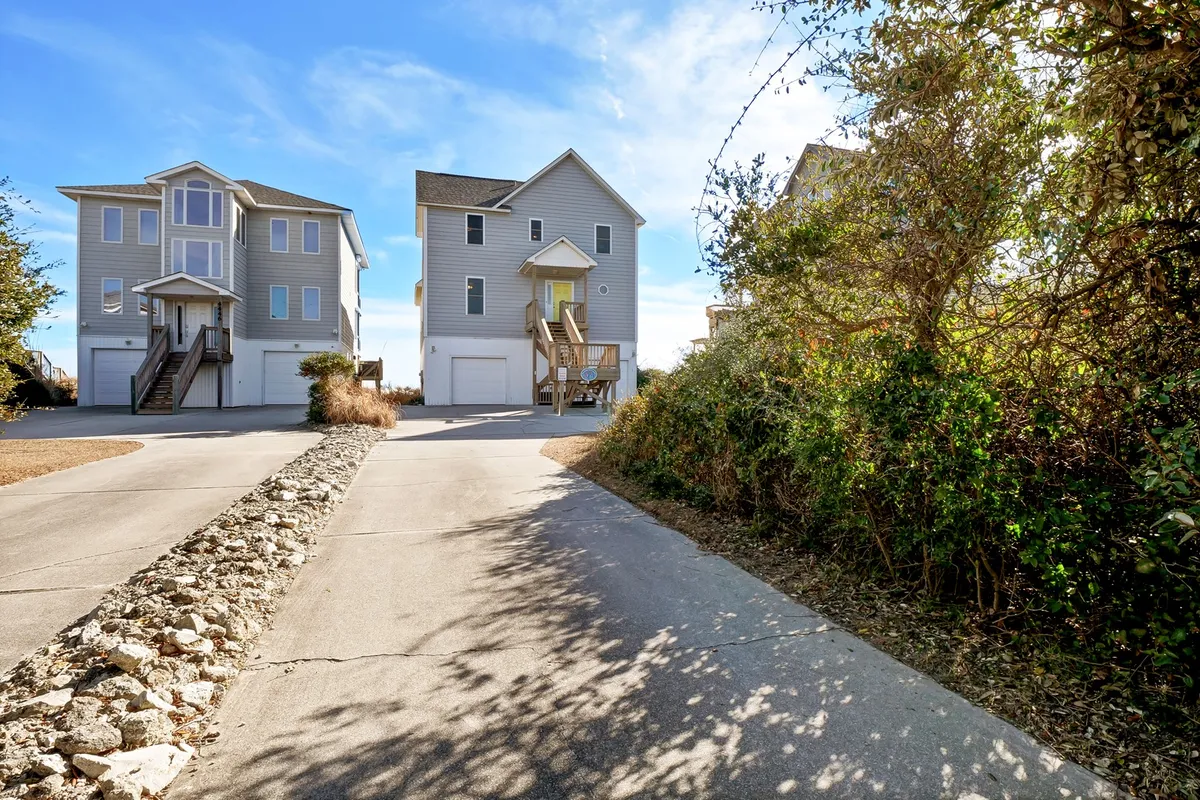 An inviting exterior view of two beach houses, with a driveway, lush greenery, and broad decks facing the ocean. The homes are positioned to enjoy sunsets, barbecues, and quiet gatherings outdoors. This image hints at the social, communal aspect of a group stay, with ample space for families or friends to gather and relax together in a beautiful coastal setting.