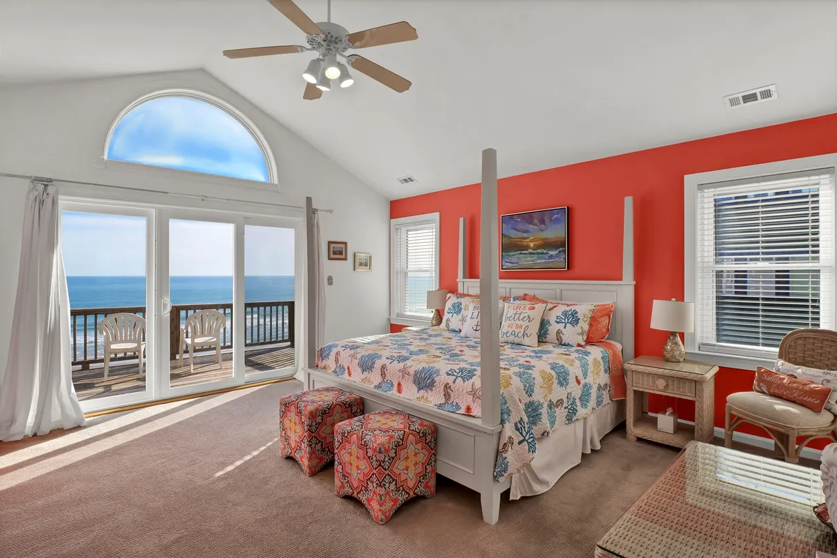 A spacious master bedroom featuring a four-poster bed with vibrant, colorful bedding. Large windows flood the room with natural light and provide stunning ocean views. Decorative stools and soft lighting create a tranquil ambiance, while the private balcony visible through the windows invites moments of relaxation and reflection.