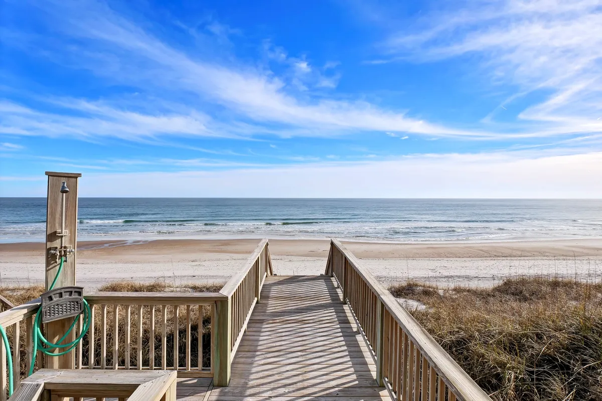 A wooden walkway leads from the property directly to the beach and ocean, flanked by a shower area, soft sand, and blue skies. The scene radiates easy accessibility with inviting beach activities moments away from home.