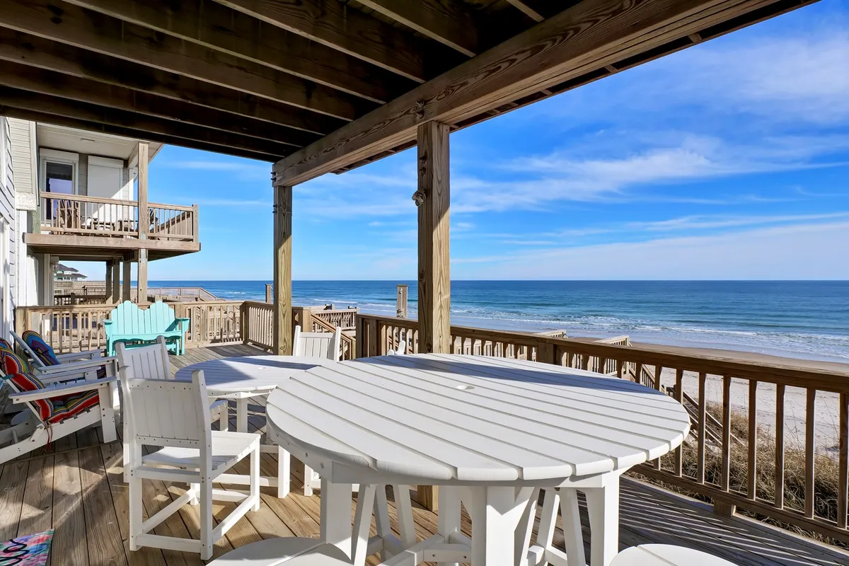 A bright, spacious wooden deck furnished with a round dining table and colorful chairs overlooks the beach and ocean. The direct sightline from the deck to the sand highlights the immediate proximity to the shore. The mood is tranquil and cheerful, perfect for enjoying meals or cocktails while taking in the ocean breeze.